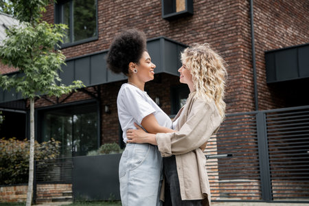 side view of interracial lesbian couple embracing and smiling at each other near new own cottageの写真素材