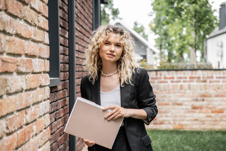 blonde and stylish property realtor holding folder and looking at camera near building on streetの写真素材