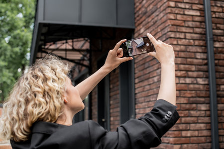 blonde real estate agent with mobile phone taking photo of modern house on city streetの写真素材
