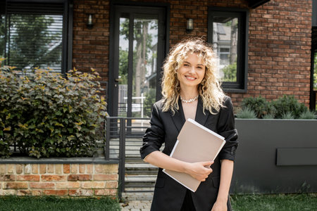cheerful blonde real estate agent holding documents and looking at camera near house on city streetの写真素材