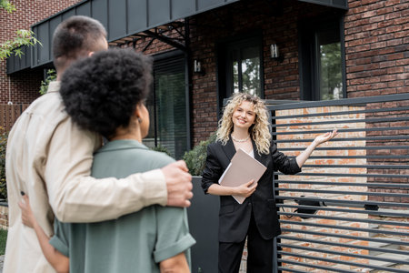 cheerful property agent with folder showing new city cottage to multiethnic couple of buyersの写真素材