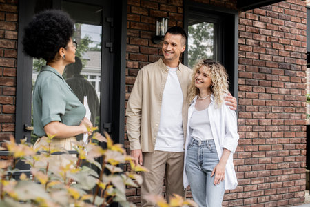 pleased couple embracing while talking with african american realtor near new cottage on streetの写真素材