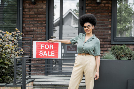 optimistic african american property agent standing with for sale signboard near fence of cottageの写真素材