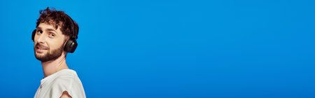 positive man in wireless headphones listening music on blue background, male fashion, audio, banner