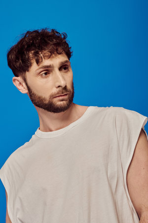 bearded and curly man posing on blue background, white tank top, male fashion, looking away