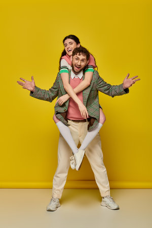 excited couple, funny, young man piggybacking brunette woman on yellow backdrop, student outfitsの写真素材