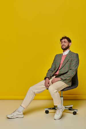 happy bearded man sitting on office chair, yellow backdrop, student in college outfit, glassesの写真素材