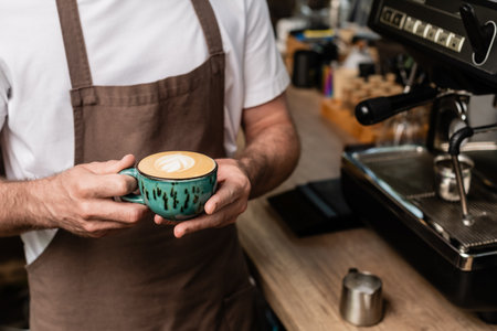 cropped view of barista in apron holding cappuccino cup while working in coffee shopの写真素材