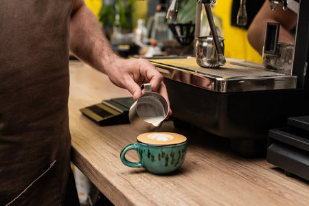 cropped view of barista in apron pouring milk from pitcher in coffee cup for cappuccino in cafeの写真素材