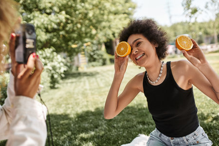 happy african american woman posing with half orange near girlfriend with vintage camera in parkの写真素材