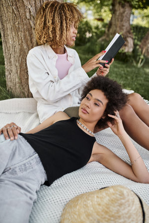 young african american woman lying near girlfriend reading book on blanket, summer leisure in parkの写真素材