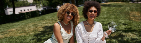 summer picnic, happy african american woman with wine glasses near stylish girlfriend, bannerの写真素材