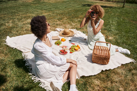 african american woman taking photo on digital camera during picnic with girlfriendの写真素材