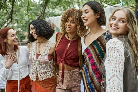 positive and trendy multiethnic women in boho clothes hugging outdoors in retreat centerの写真素材