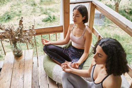 young woman meditating in lotus pose near multiracial girlfriend in patio of retreat centerの写真素材
