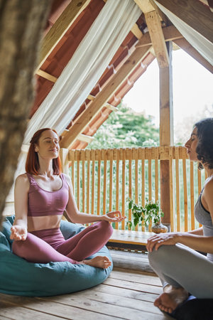 smiling woman with closed eyes sitting in lotus pose near multiracial girlfriend in cozy patioの写真素材