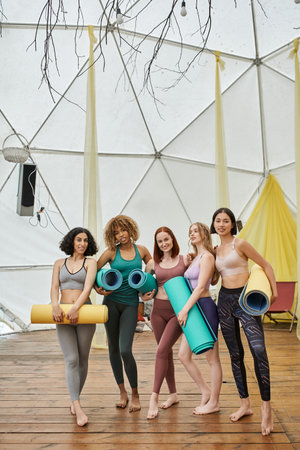 full length of multiethnic women in sportswear standing with yoga mats in modern retreat centerの写真素材