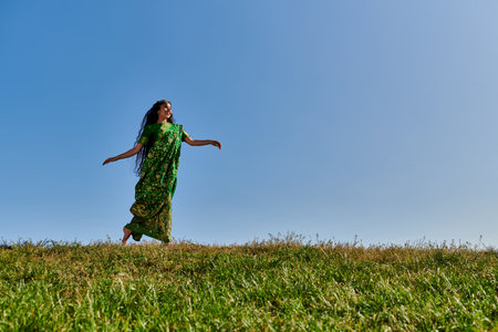 vibrant summer, joyful indian woman in ethnic clothes running in green field under blue skyの写真素材
