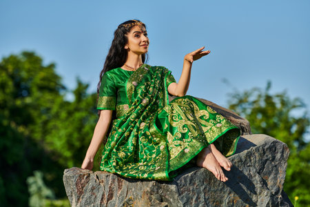 smiling and stylish barefoot indian woman in sari sitting on stone with blue sky on background
