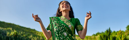 happy young indian woman in green sari standing with summer landscape on background, bannerの写真素材