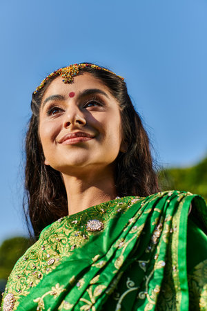 low angle view of cheerful indian woman with bindi and sari standing with sky on background