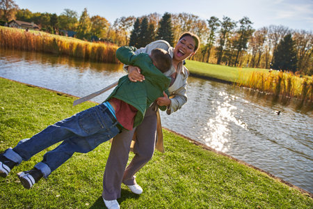 playful, joy, happy african american mother lifting son, having fun near pond, outerwear, autumnの写真素材