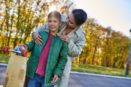 african american woman hugging son with penny board, autumn, fall season, motherly love, parkの写真素材