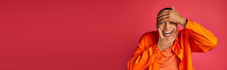 playful african american man looking at camera and touching face, red background, banner, copy space
