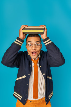 astonished african american student in trendy jacket and eyeglasses with notebooks on head on blueの写真素材