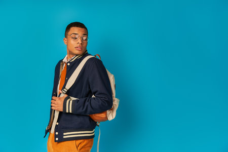 trendy african american student in eyeglasses and jacket looking at backpack on blue backgroundの写真素材