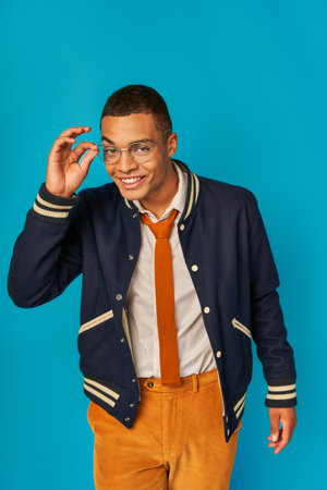 charismatic african american student in jacket adjusting eyeglasses and smiling at camera on blueの写真素材
