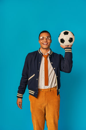 happy african american student in jacket and orange pants playing with soccer ball on blueの写真素材