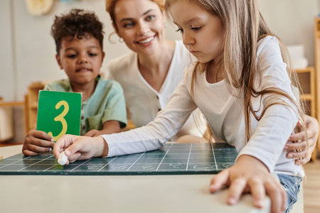 girl writing on chalkboard near african american boy with number and teacher, Montessori school