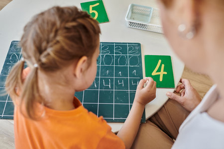 top view of clever girl writing on chalkboard, learning how to count near teacher, Montessori school