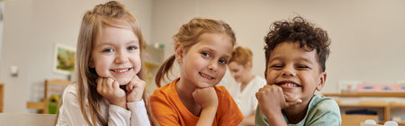smiling and interracial kids looking at camera in blurred class in montessori school, banner