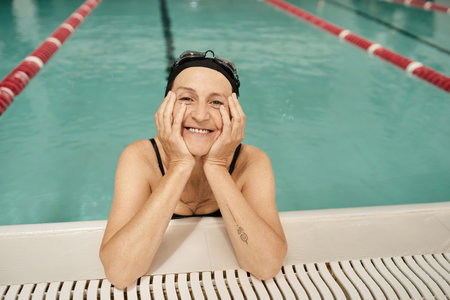 joyous middle aged woman in swim cap and goggles looking at camera, water in pool, recreation centerの写真素材