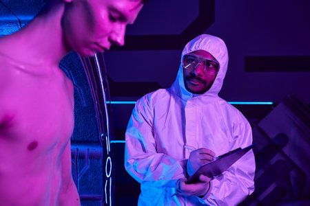 indian scientist in hazmat suit writing on clipboard, examining humanoid alien in discovery centerの写真素材