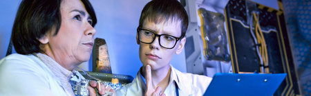 serious young intern looking at middle aged woman scientist in futuristic discovery center, banner