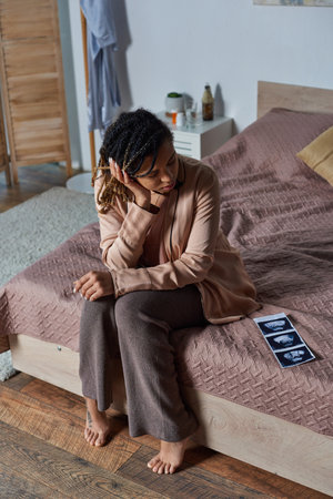 african american woman sitting on bed and looking at ultrasound photo, glass of water, birth control