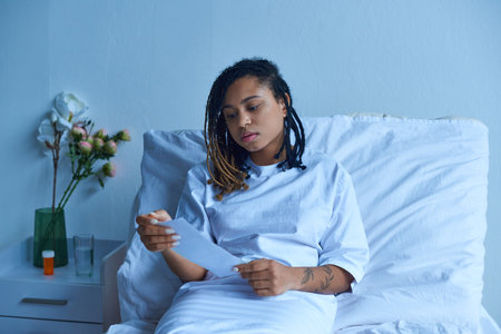 private ward, sad african american woman in hospital gown looking at ultrasound, miscarriage conceptの写真素材