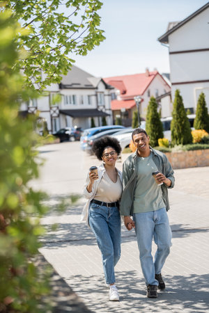 smiling african american couple with coffee to go looking at camera near houses on urban streetの写真素材