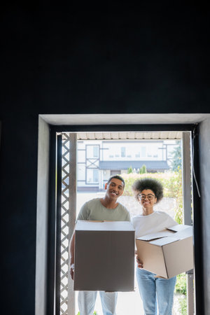 smiling african american couple holding carton boxes and looking at camera near door in new house