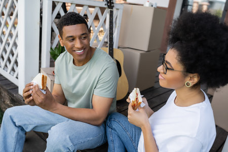 smiling african american man holding sandwich near girlfriend and boxes on porch of new houseの写真素材