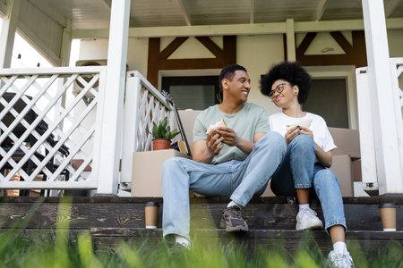 smiling african american couple holding sandwiches near carton boxes on porch of new houseの写真素材
