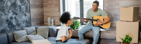 happy african american couple with coffee playing acoustic guitar near packages in new house, banner