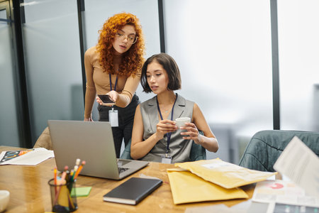 redhead businesswoman with smartphone near colleague with coffee cup working on laptop in office