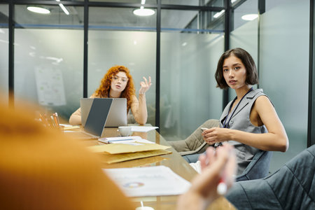young businesswomen looking at blurred colleague near laptops and documents in meeting room