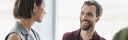 joyful bearded entrepreneur smiling near young businesswoman in modern office, banner