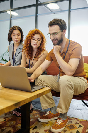teamwork, smiling bearded entrepreneur looking at laptop near smiling businesswomen in modern officeの写真素材