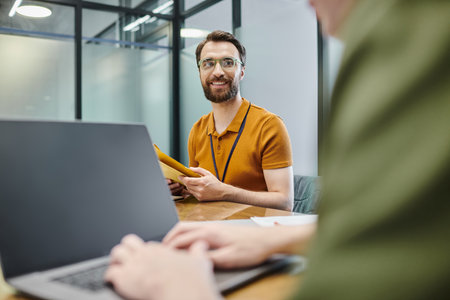 bearded businessman with envelopes smiling near blurred colleague typing on laptop in office